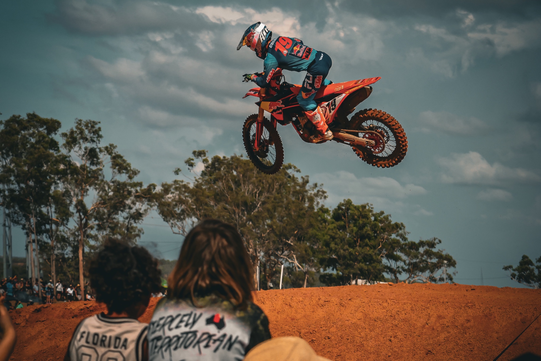 Motocross rider soaring over a jump with spectators watching from the foreground.